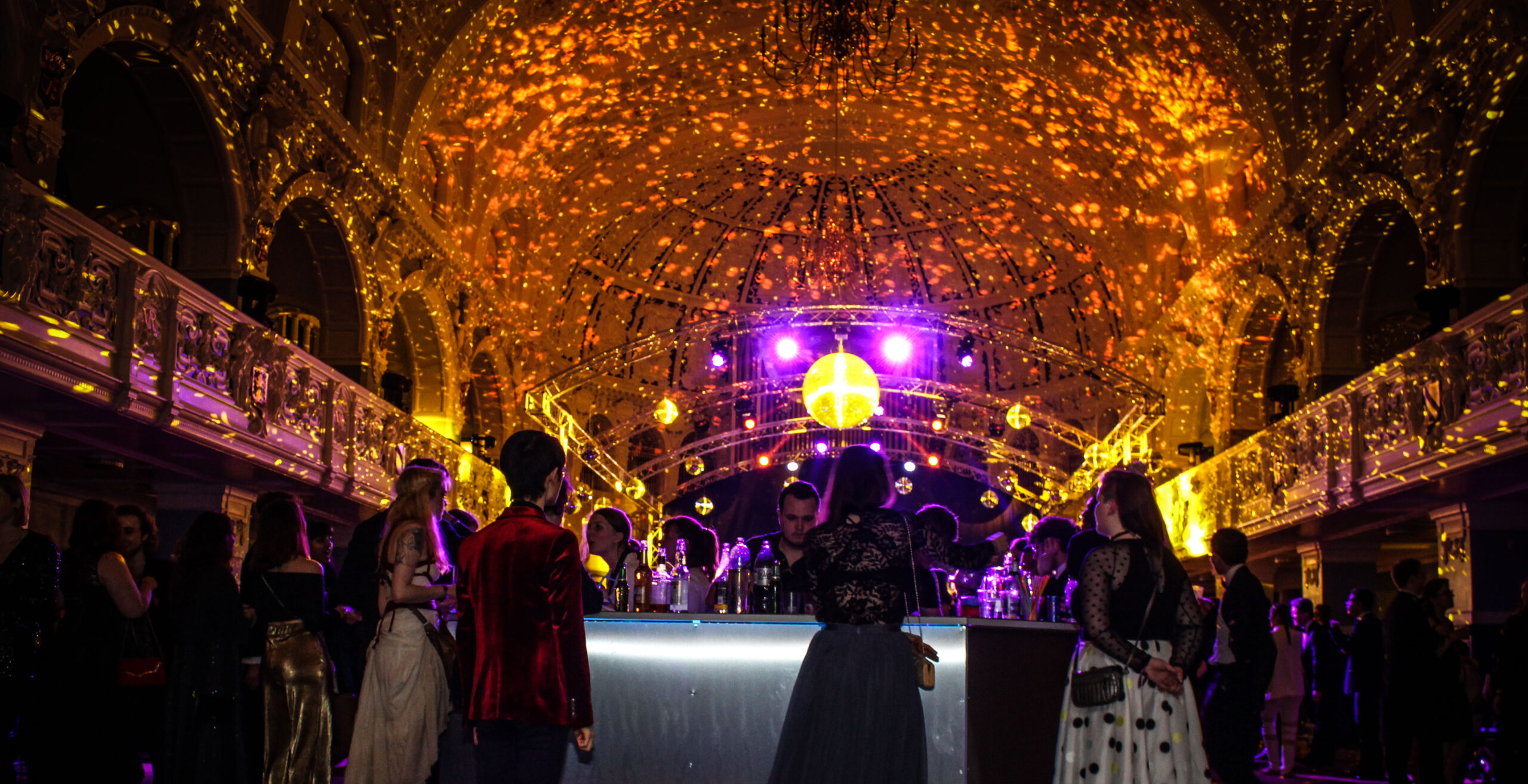 Image from Glitterball 2017. People standing at bar, bathed in orange glow from the lights